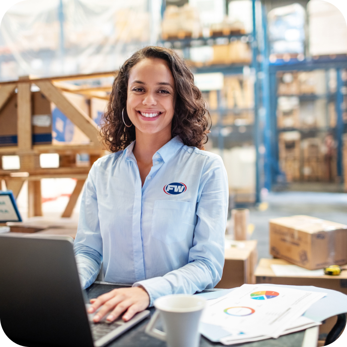 women in a warehouse wearing a blue button up with FW logo her left side smiling into the camera while on a laptop.